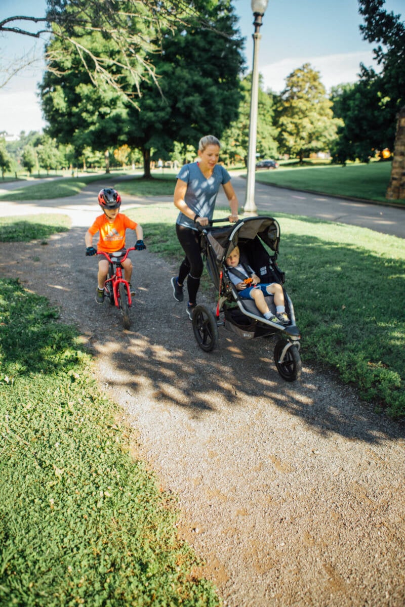 a woman pushing a stroller and teaching her child to ride a bike while running