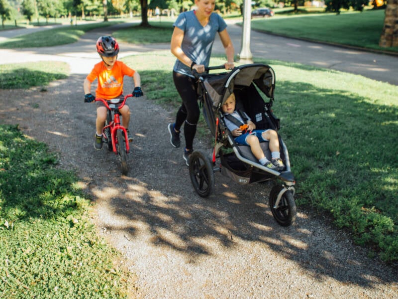 a woman pushing a stroller and teaching her child to ride a bike while running